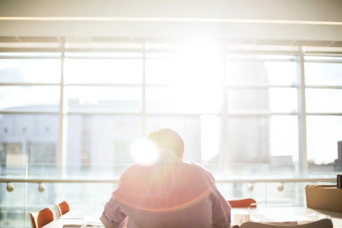 Man sitting at a desk with his back to the camera, facing a window with the sun shining through.