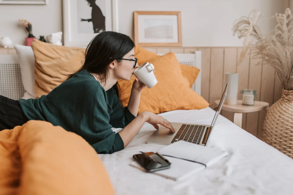 Woman working on laptop in bed while sipping from a mug.