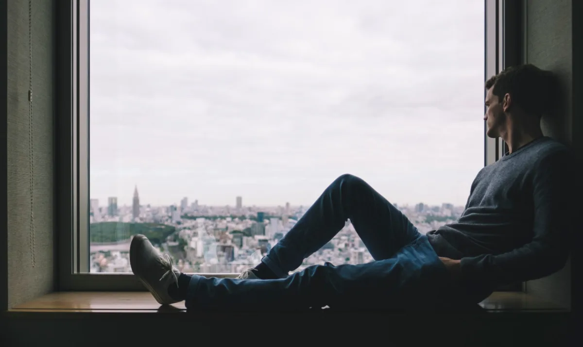 A man sitting by a large window with a view of the city skyline, looking contemplatively into the distance.