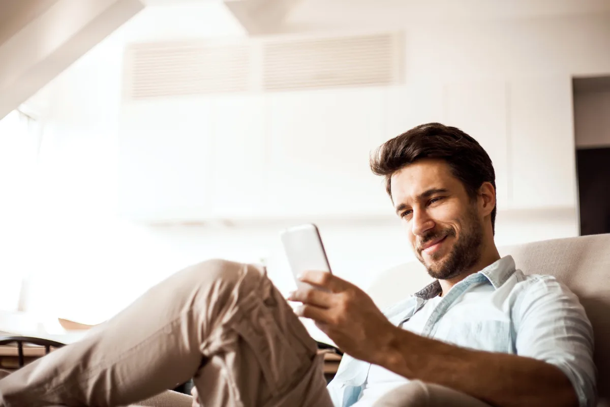 Man smiling while looking at his smartphone, comfortably seated on a couch.