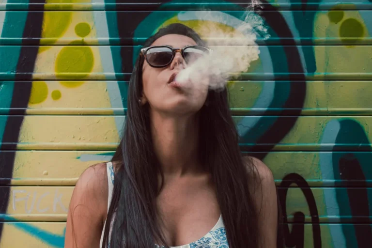 Woman exhaling smoke against a graffiti-covered wall.