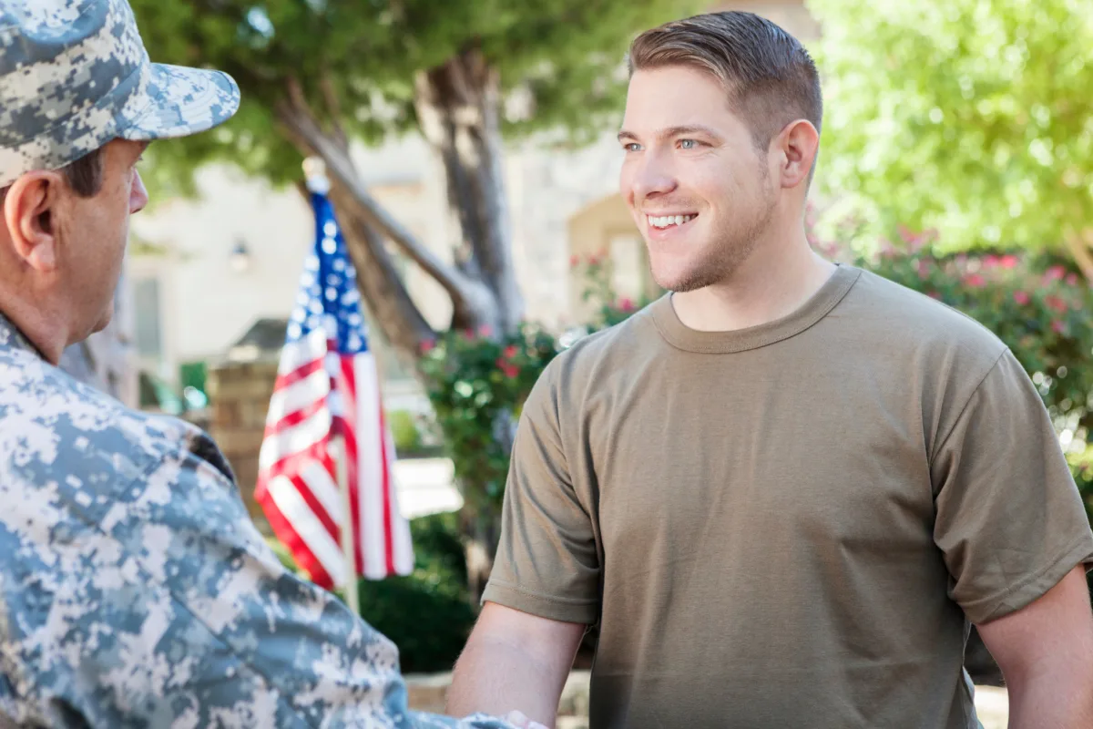 A civilian man smiling at a person in military uniform with an american flag in the background.