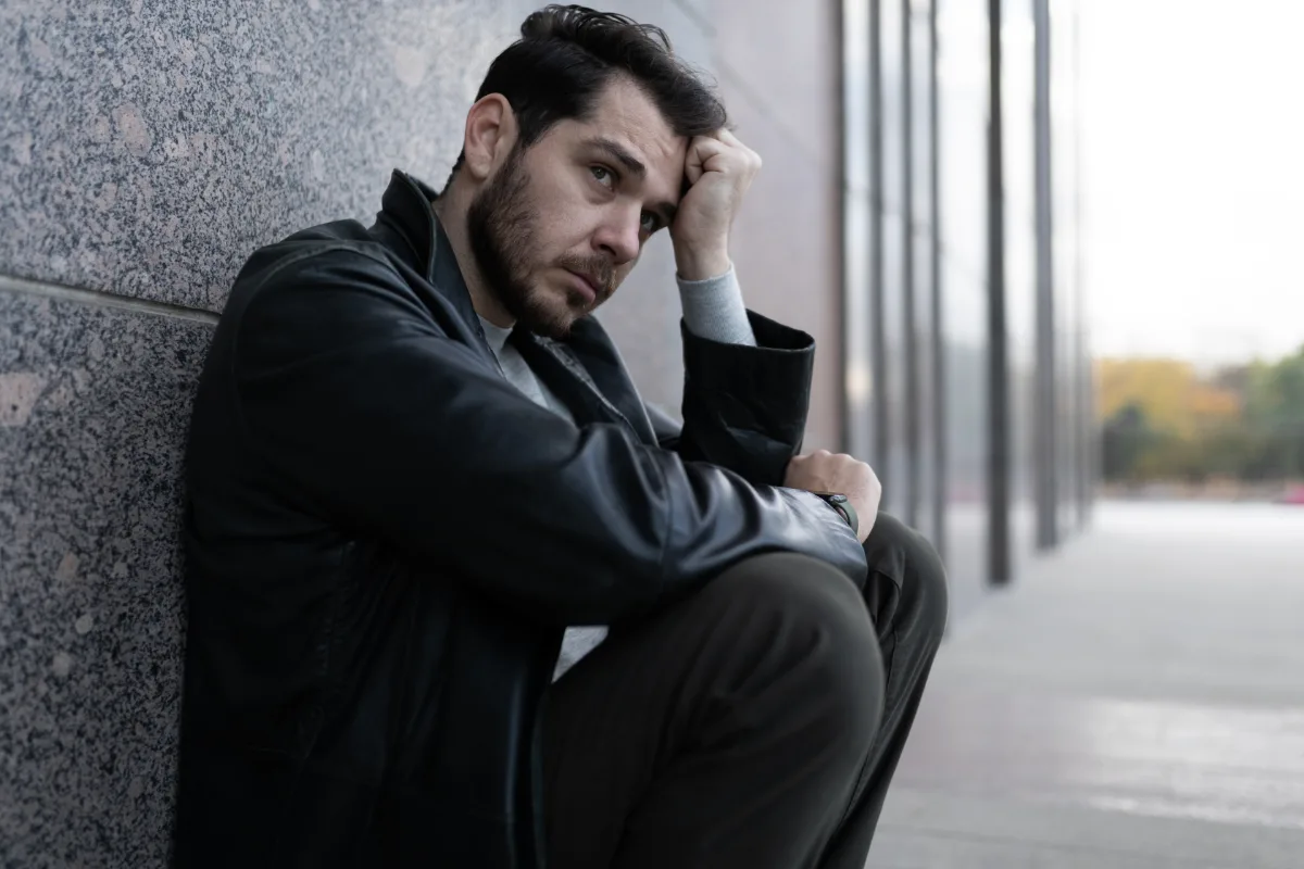 Man sitting on the ground against a wall, appearing pensive or troubled.