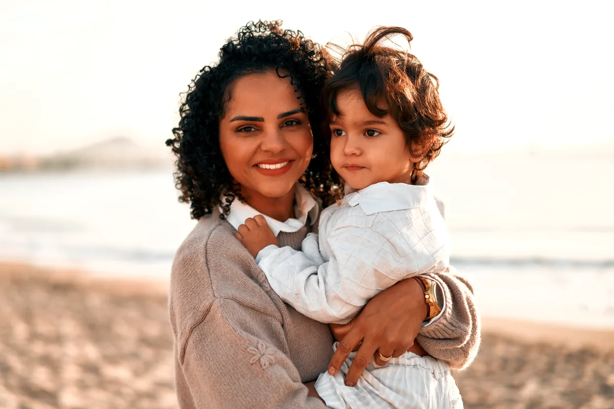 A woman holding a young child at the beach during sunset.