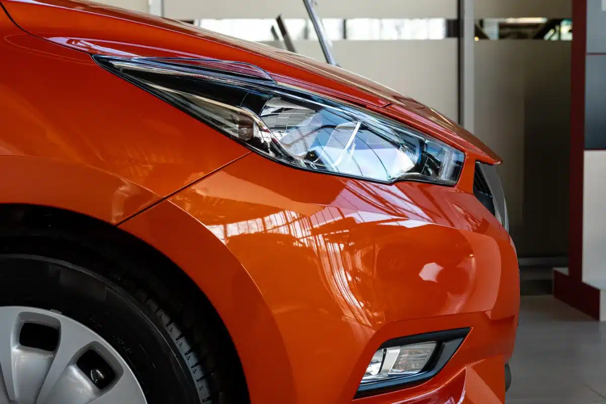 A close up of an orange car in a showroom, with the driver cautiously examining the vehicle's features.