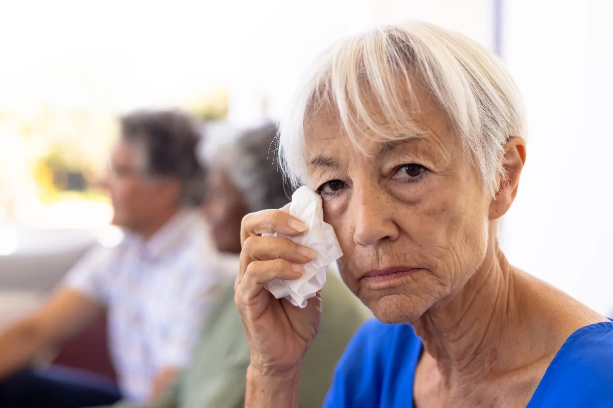 Senior woman holding a tissue, appearing concerned or unwell, with other people in the background.