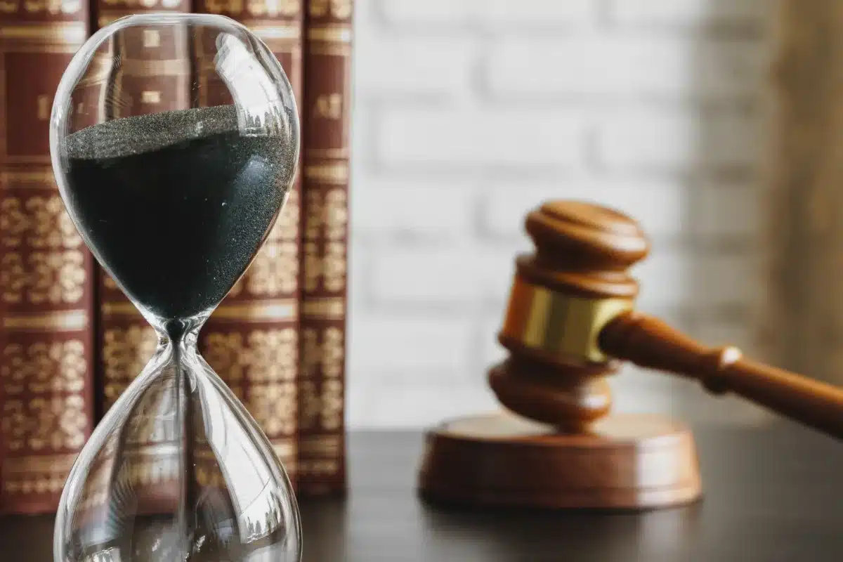 An hourglass with sand next to a gavel on a table, symbolizing the passage of time in legal proceedings. The presence of the gavel suggests the authority and power in courtrooms