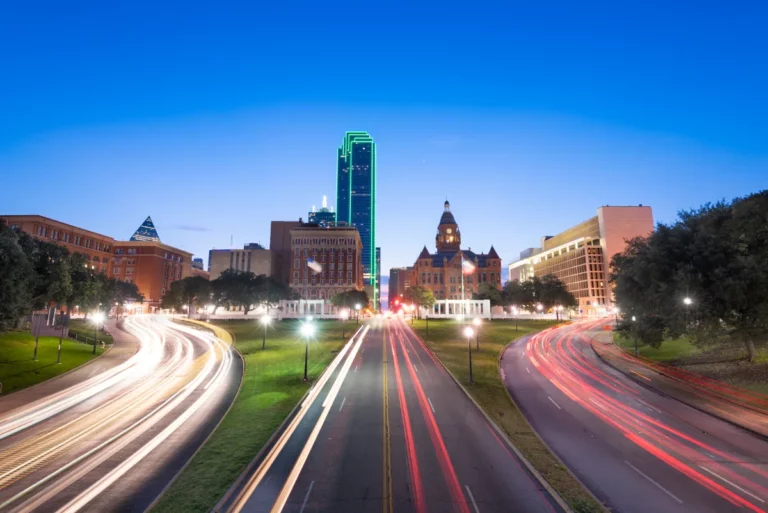 Twilight cityscape with light trails from traffic on a bustling road in Texas, flanked by illuminated modern and historic architecture.