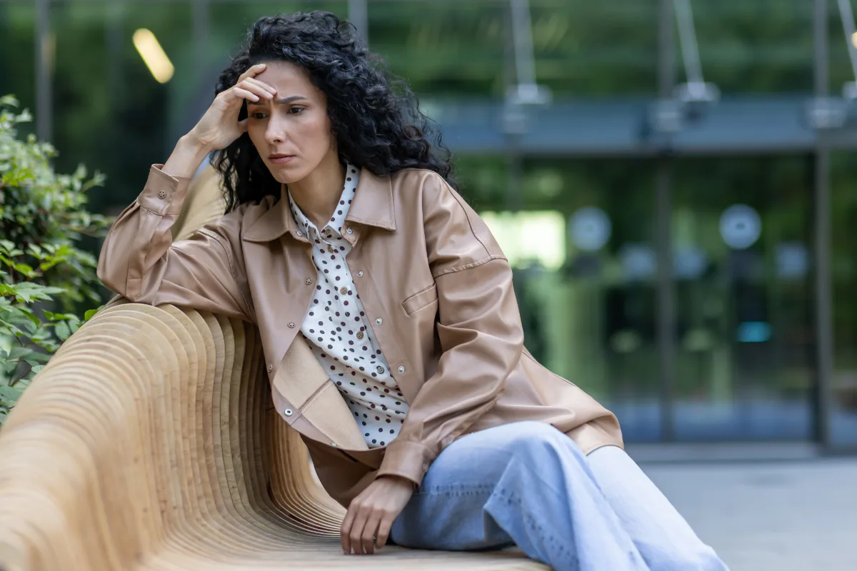 A pensive woman sitting on an outdoor bench with a hand to her forehead.