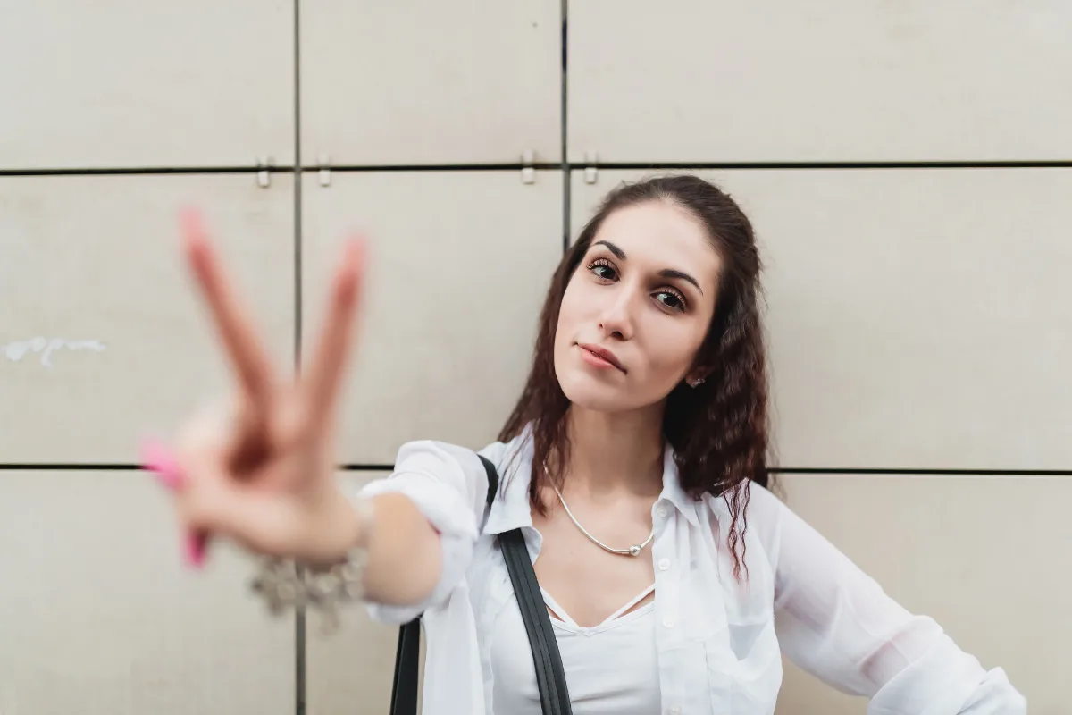 A woman gesturing a peace sign towards the camera with a neutral background.