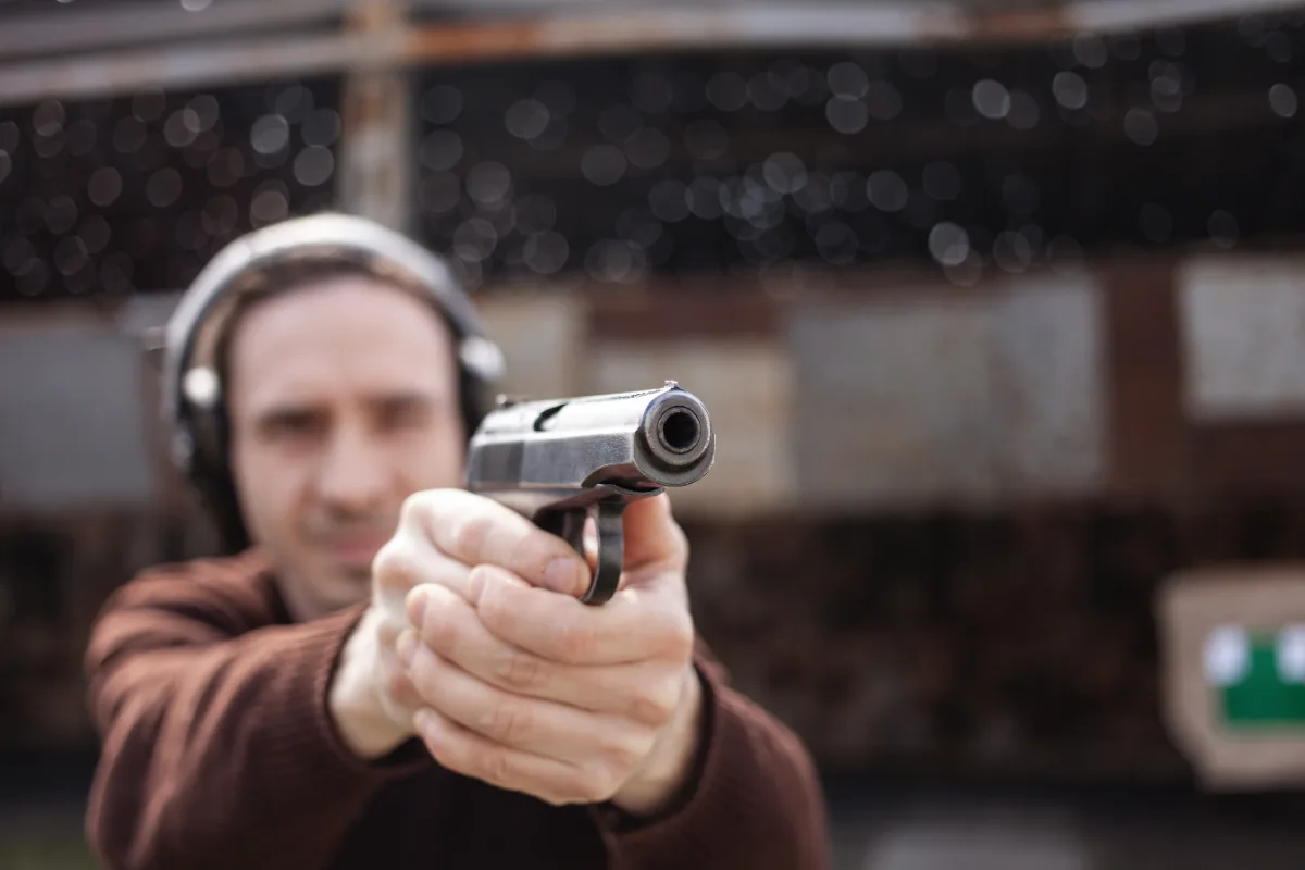 Man aiming a handgun directly at camera with focus on the muzzle, wearing protective ear muffs.