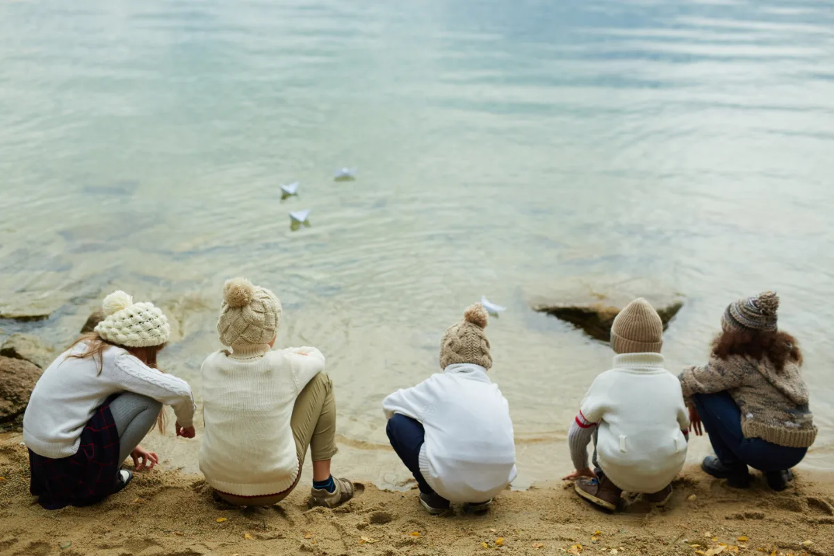 A group of children sitting on the sand next to a body of water charges.