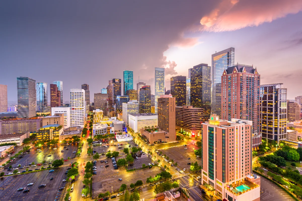 A vibrant cityscape at dusk with illuminated skyscrapers, dramatic clouds, and the notable presence of a Texas Assault Lawyer billboard.