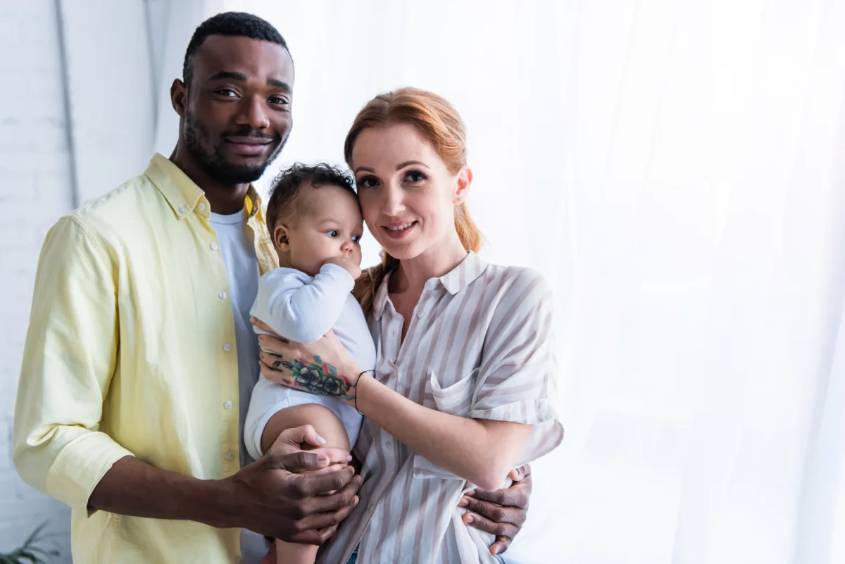 A multiracial couple holding their baby with affectionate smiles.