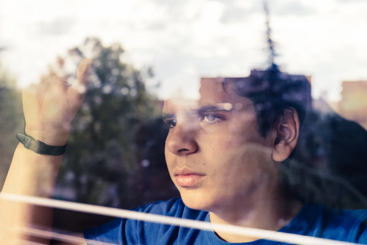 A young man gazing outside through a window with reflections of trees overlaying his thoughtful expression.