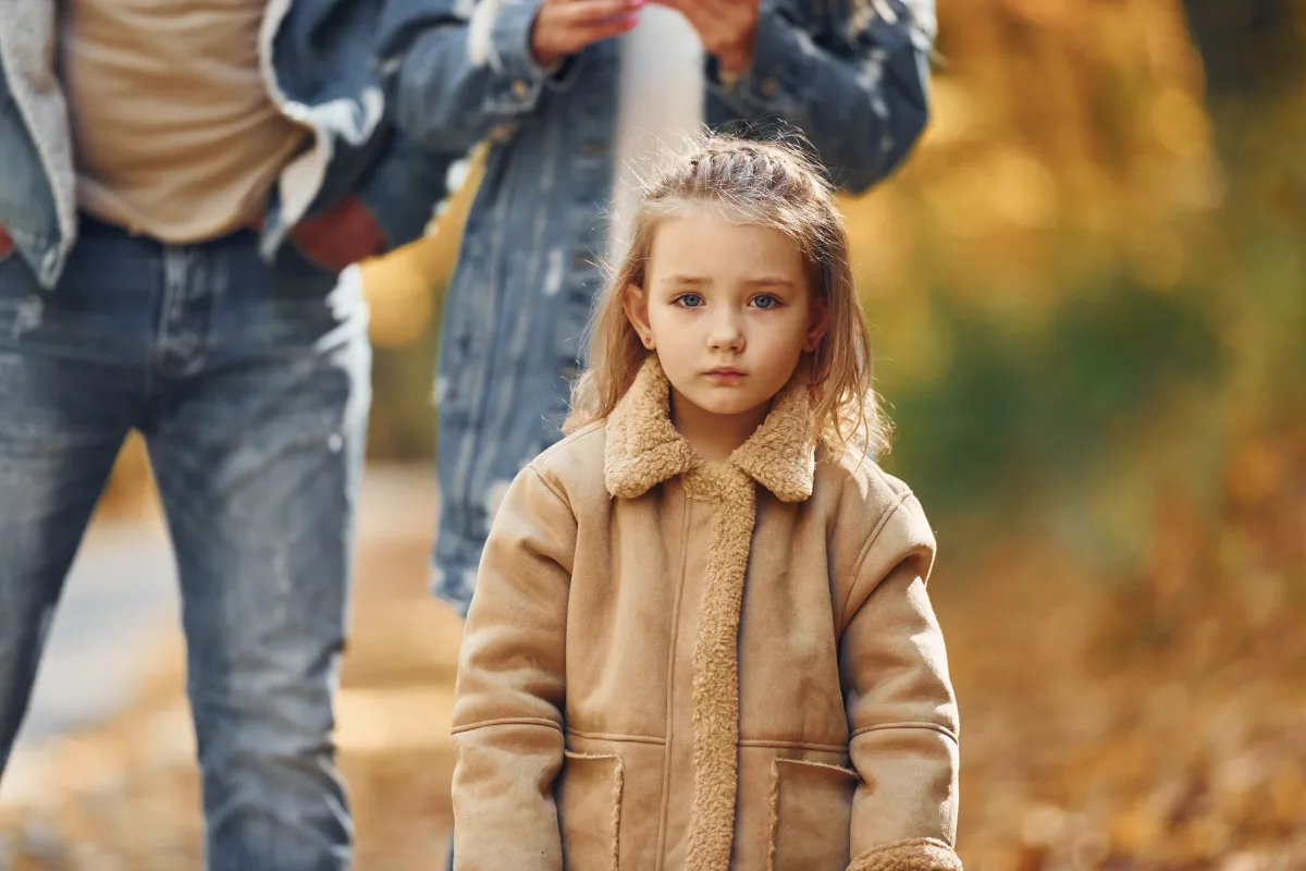 Young girl in a coat standing in front of two adults during autumn.