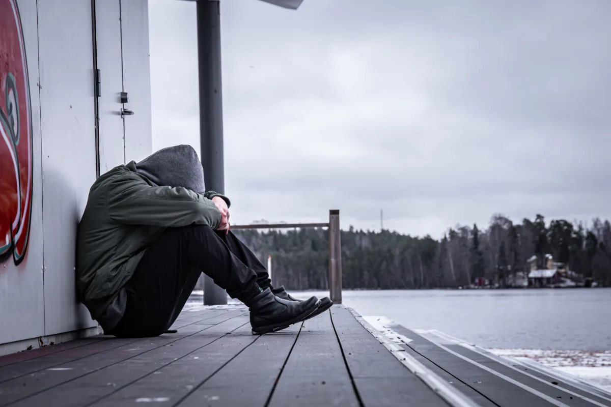 In this scene, a man wearing a hoodie peacefully sits on a dock by the serene setting of a lake.