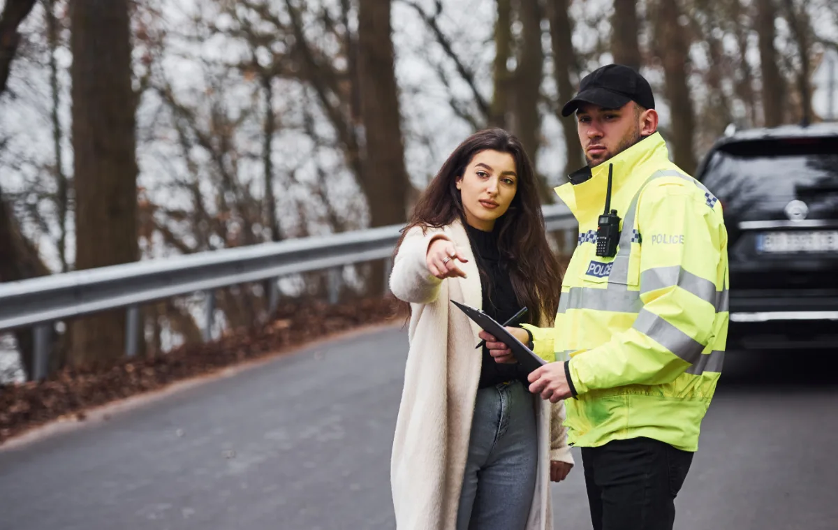 A police officer in a reflective vest talks to a civilian woman by the roadside.