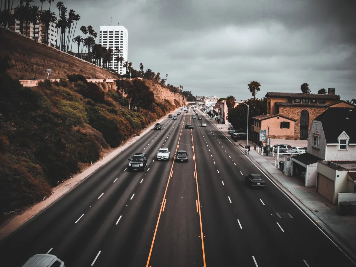 Vehicles driving on a multi-lane highway with palm trees and buildings alongside under an overcast sky.