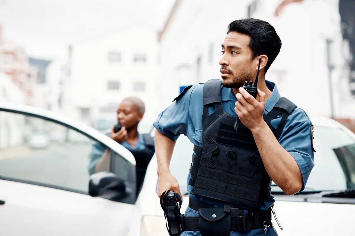 A security officer communicating via radio while standing near a vehicle, with another officer in the background.