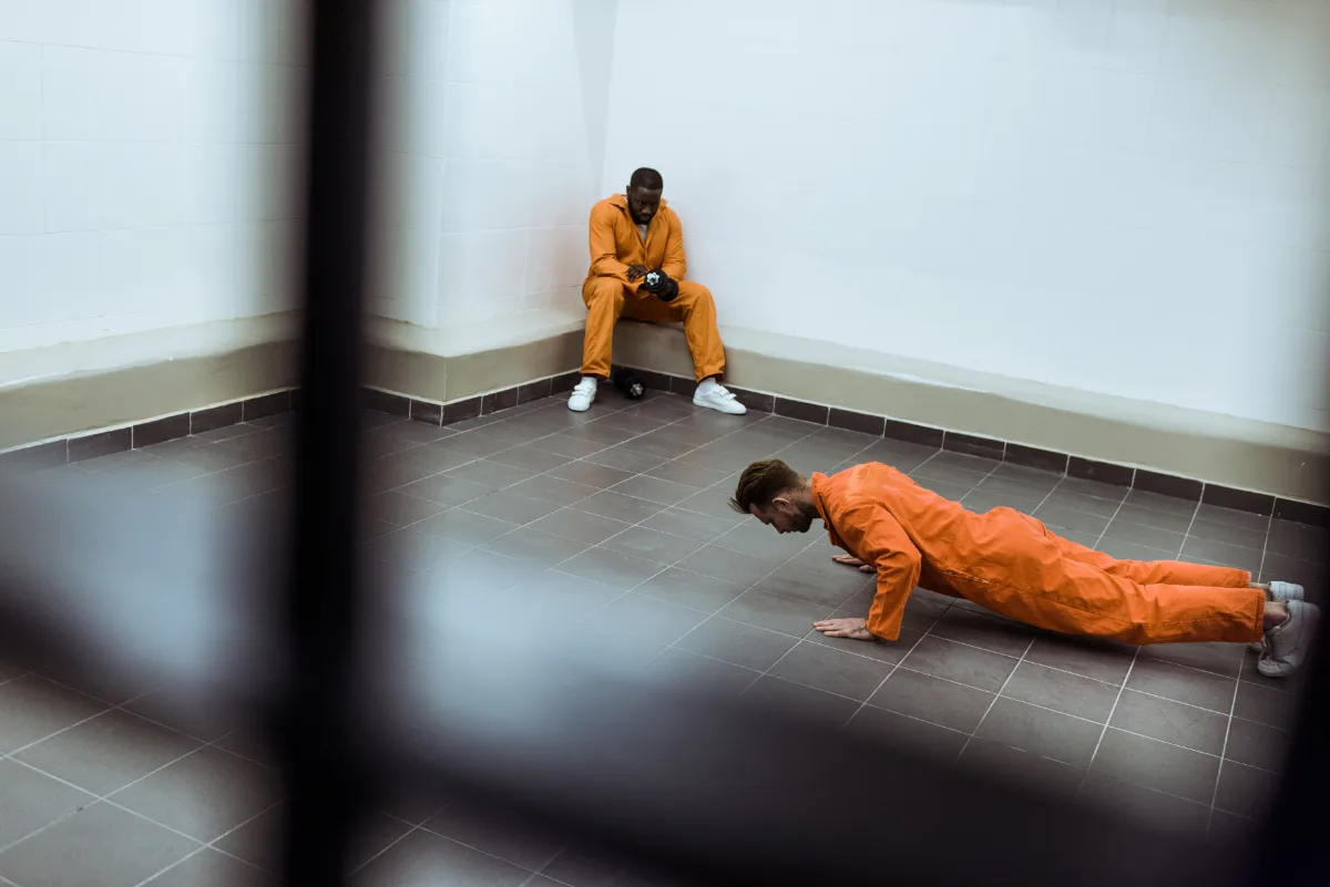 Two individuals in orange jumpsuits in a room, one doing push-ups and the other sitting against the wall.