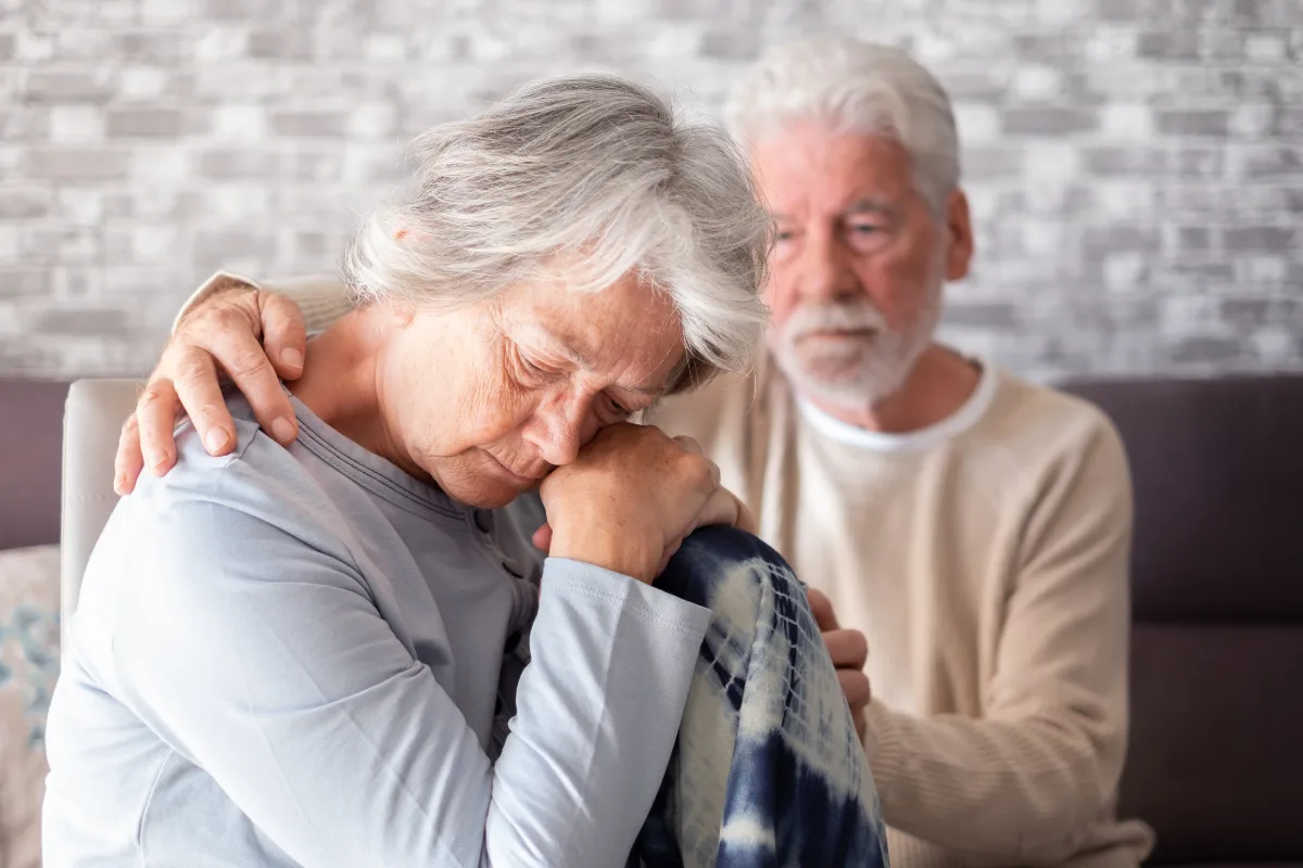 An elderly woman appears distressed and is being comforted by an elderly man.