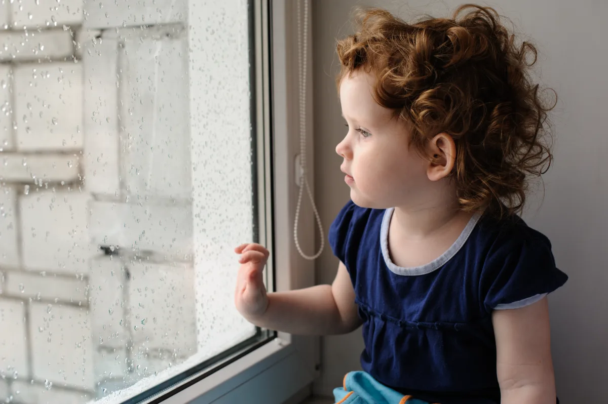 A young child with curly hair looking out a rain-spattered window.
