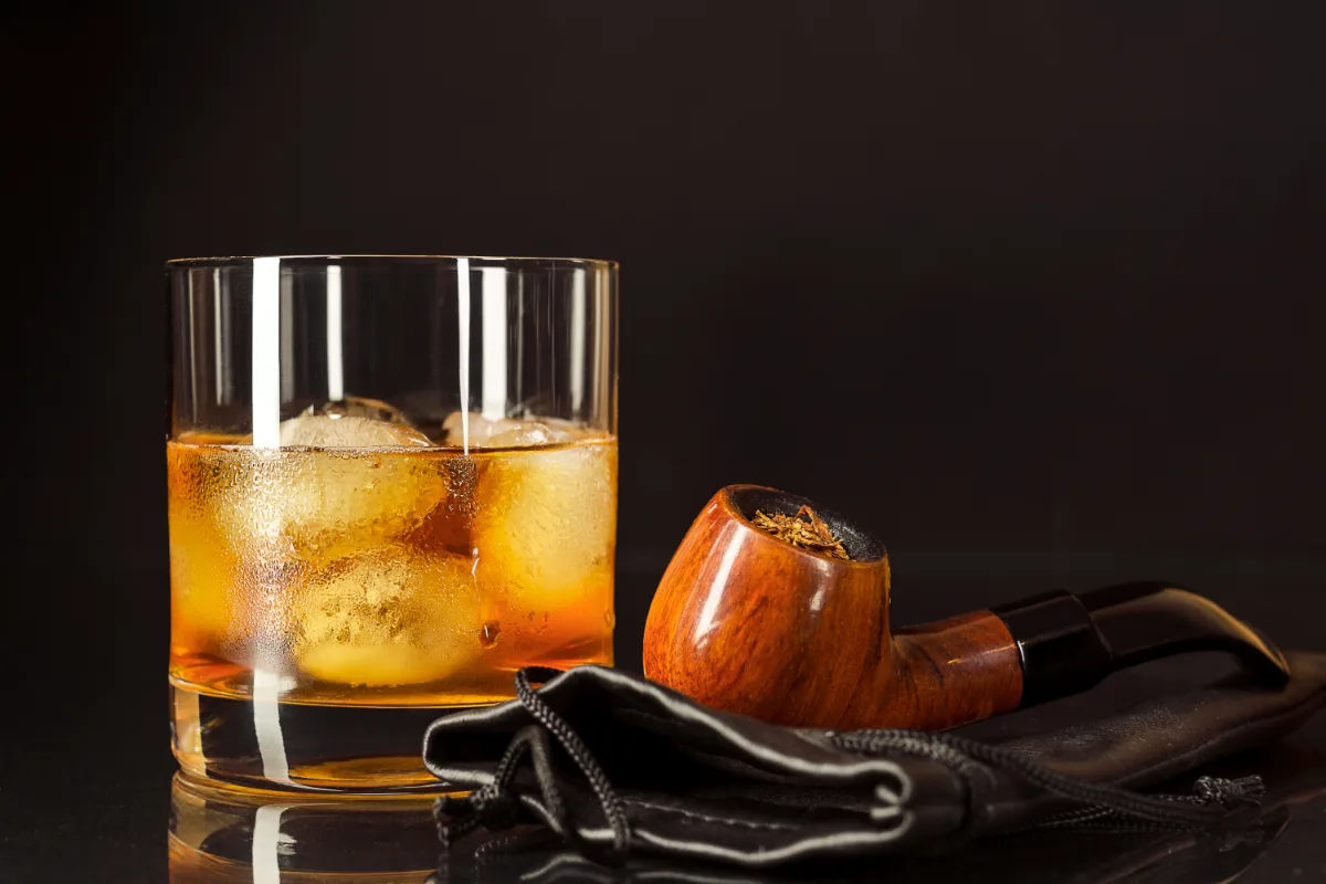 A glass of whiskey with ice and a pipe on a black background, creating a constitutional mood.