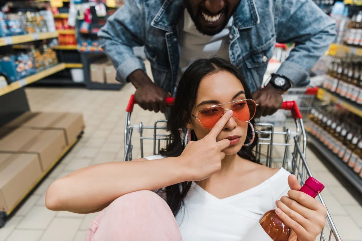 A woman wearing sunglasses sits in a shopping cart, playfully posing with a peace sign, while a man behind her smiles and pushes the cart in a supermarket aisle.