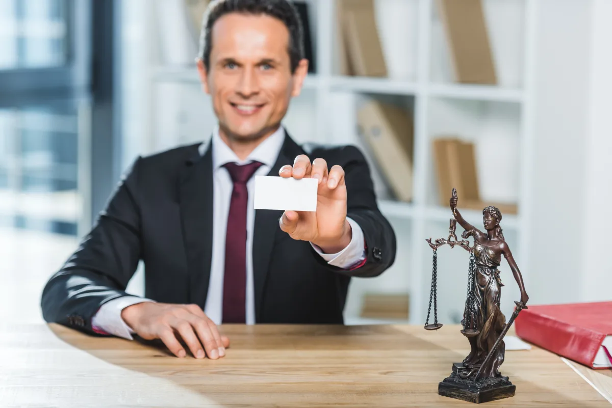 A businessman holding a blank business card with a statue of lady justice on his desk.
