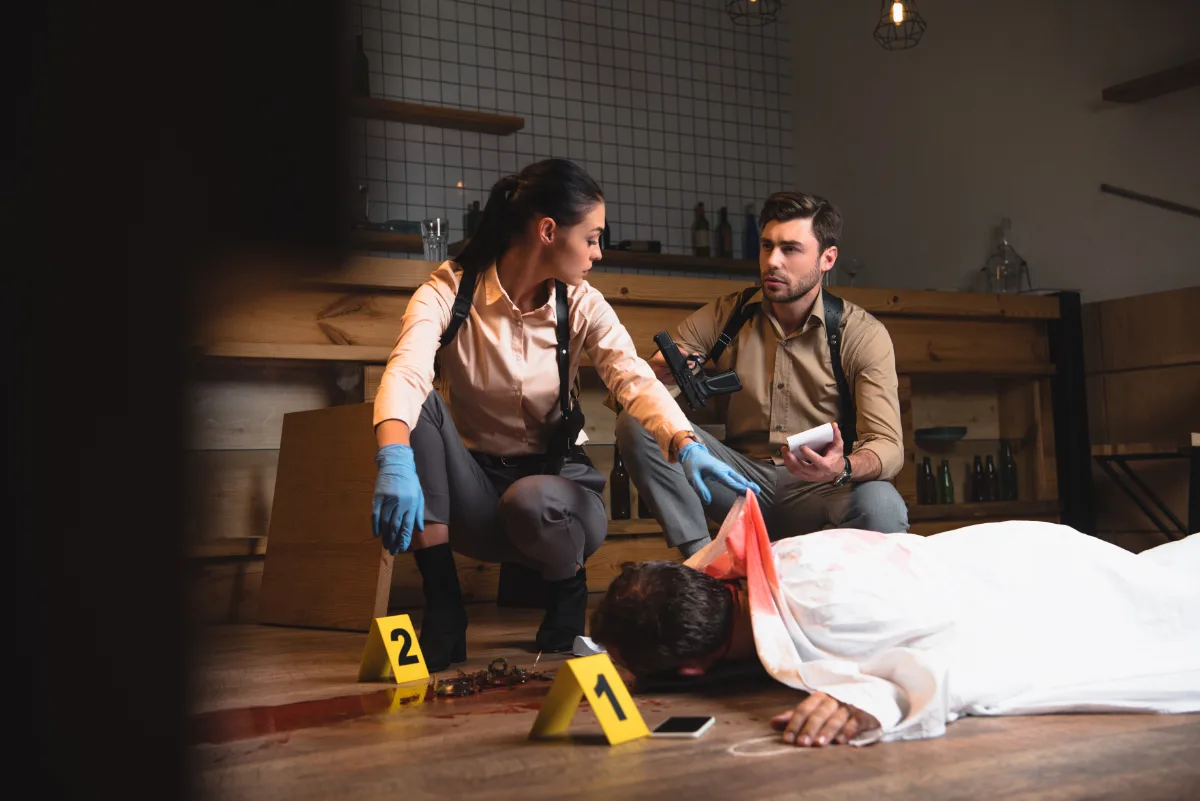 Two crime scene investigators examining evidence near a body at a crime scene.