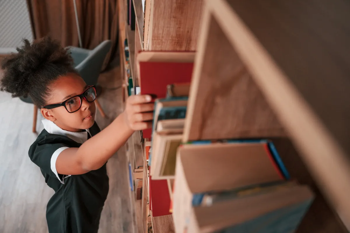 A girl in glasses is reaching for a book on a shelf while wearing her probation ankle monitor.