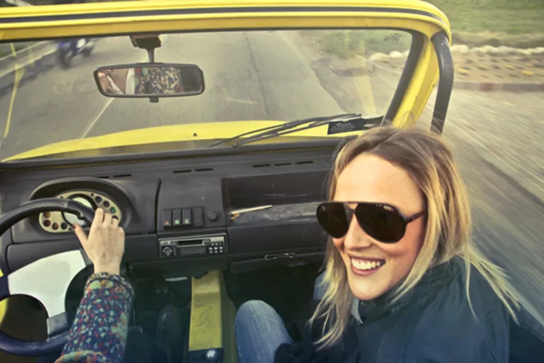 Woman wearing sunglasses driving a yellow vehicle with a road visible in the rearview mirror.