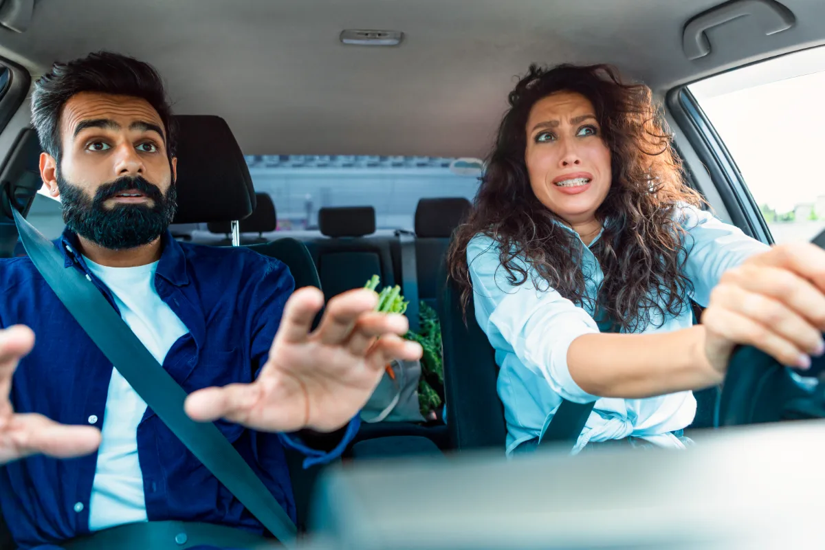 A woman driving a car appears alarmed while a male passenger looks on with an expression of concern.