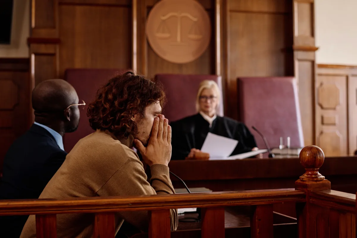 A distressed individual sits beside their lawyer in a courtroom while a judge presides over the case.
