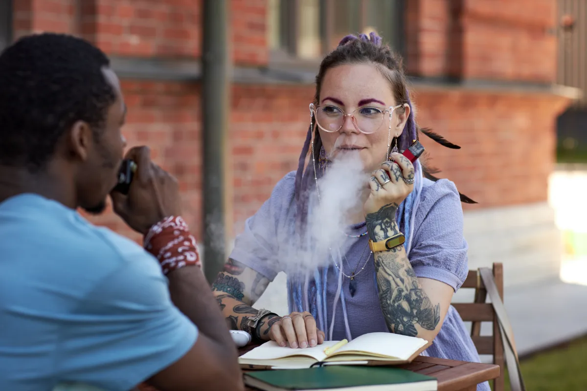 Woman exhaling vapor from an electronic cigarette while sitting at a table with a notebook and conversing with a man.