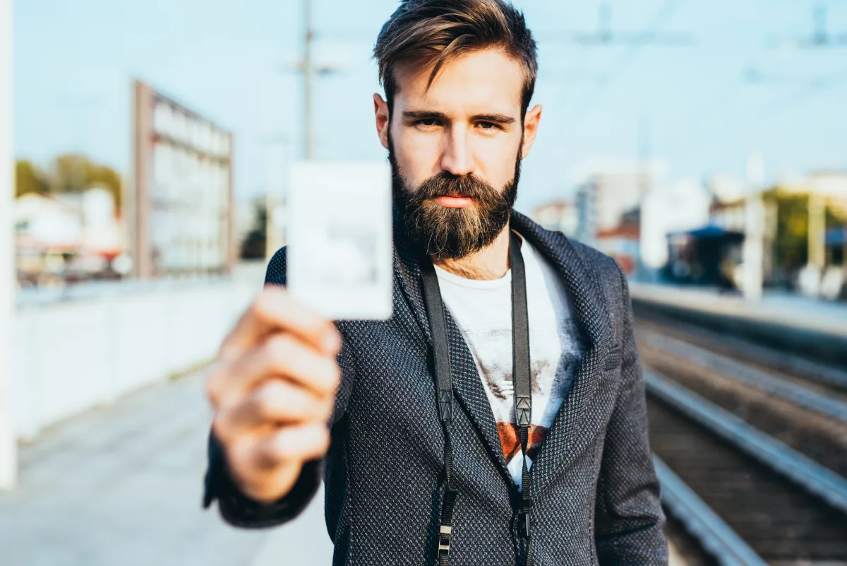A bearded man holding out a train ticket at a railway station.