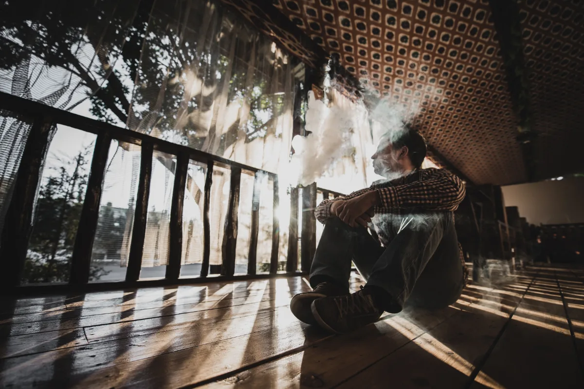 A man sitting on a wooden floor smoking a cigarette while facing misdemeanor charges.